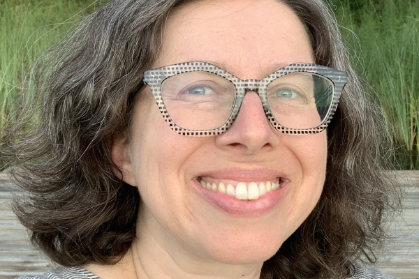 A person with curly, shoulder-length hair smiles at the camera, wearing clear, patterned glasses and a striped shirt. The background features greenery and a wooden surface