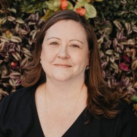 A woman with long brown hair and a slight smile stands in front of a backdrop of green and purple foliage with small red and yellow flowers. She is wearing a black top and has earrings