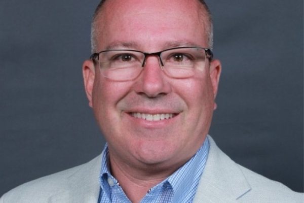 A man with short, light-colored hair and glasses smiles at the camera. He is wearing a light gray blazer over a blue checkered shirt. The background is a solid dark color