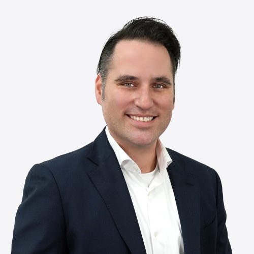 A man with dark hair styled to the side is smiling. He is wearing a dark blazer over a white shirt. The background is plain and light-colored