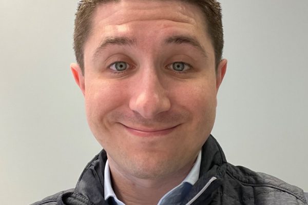A man with short brown hair and green eyes smiles at the camera. He is wearing a dark vest over a light blue shirt and is positioned against a plain light-colored background