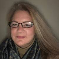 A woman with long, straight hair and glasses smiles at the camera. She is wearing a dark scarf over a light-colored top. The background is neutral and softly lit