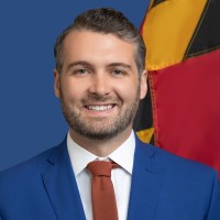 A man with short, light brown hair and a beard smiles while wearing a blue suit and an orange tie. He is positioned in front of a background featuring a flag with red, yellow, and black colors