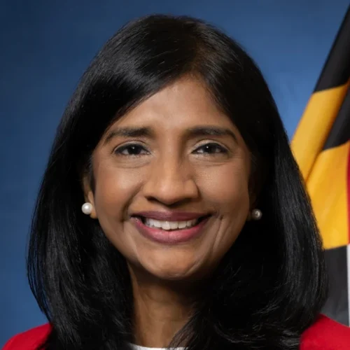 A woman with long black hair and wearing a red blazer smiles at the camera. She has pearl earrings and is positioned in front of a blue background with a flag featuring black, yellow, and red colors