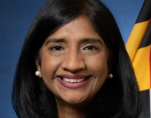 A woman with long black hair and wearing a red blazer smiles at the camera. She has pearl earrings and is positioned in front of a blue background with a flag featuring black, yellow, and red colors
