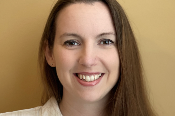 A woman with long brown hair smiles at the camera, wearing a white button-up shirt. The background is a warm, solid yellow color
