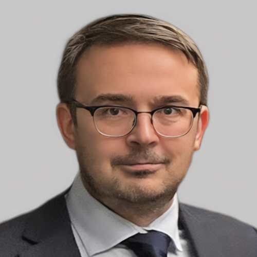 A man with short brown hair and glasses is wearing a dark suit and a light blue shirt with a tie. He is looking directly at the camera with a neutral expression against a plain gray background