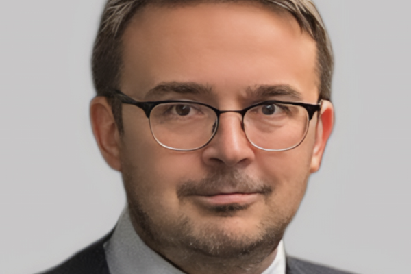A man with short brown hair and glasses is wearing a dark suit and a light blue shirt with a tie. He is looking directly at the camera with a neutral expression against a plain gray background