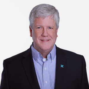 A man with gray hair and a friendly smile is wearing a dark blazer over a light blue collared shirt. He has a small blue ribbon pinned to his blazer. The background is plain and white