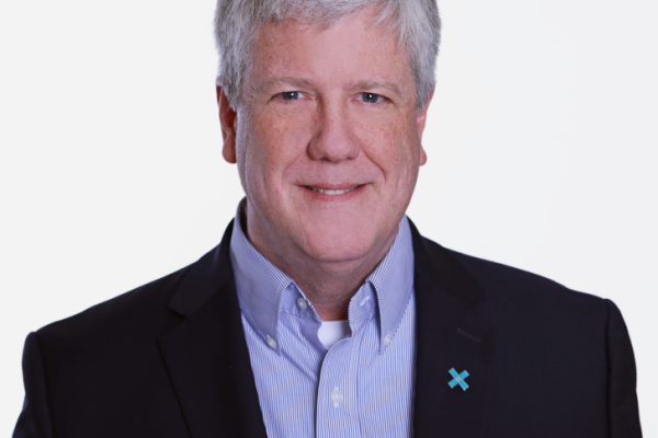 A man with gray hair and a friendly smile is wearing a dark blazer over a light blue collared shirt. He has a small blue ribbon pinned to his blazer. The background is plain and white